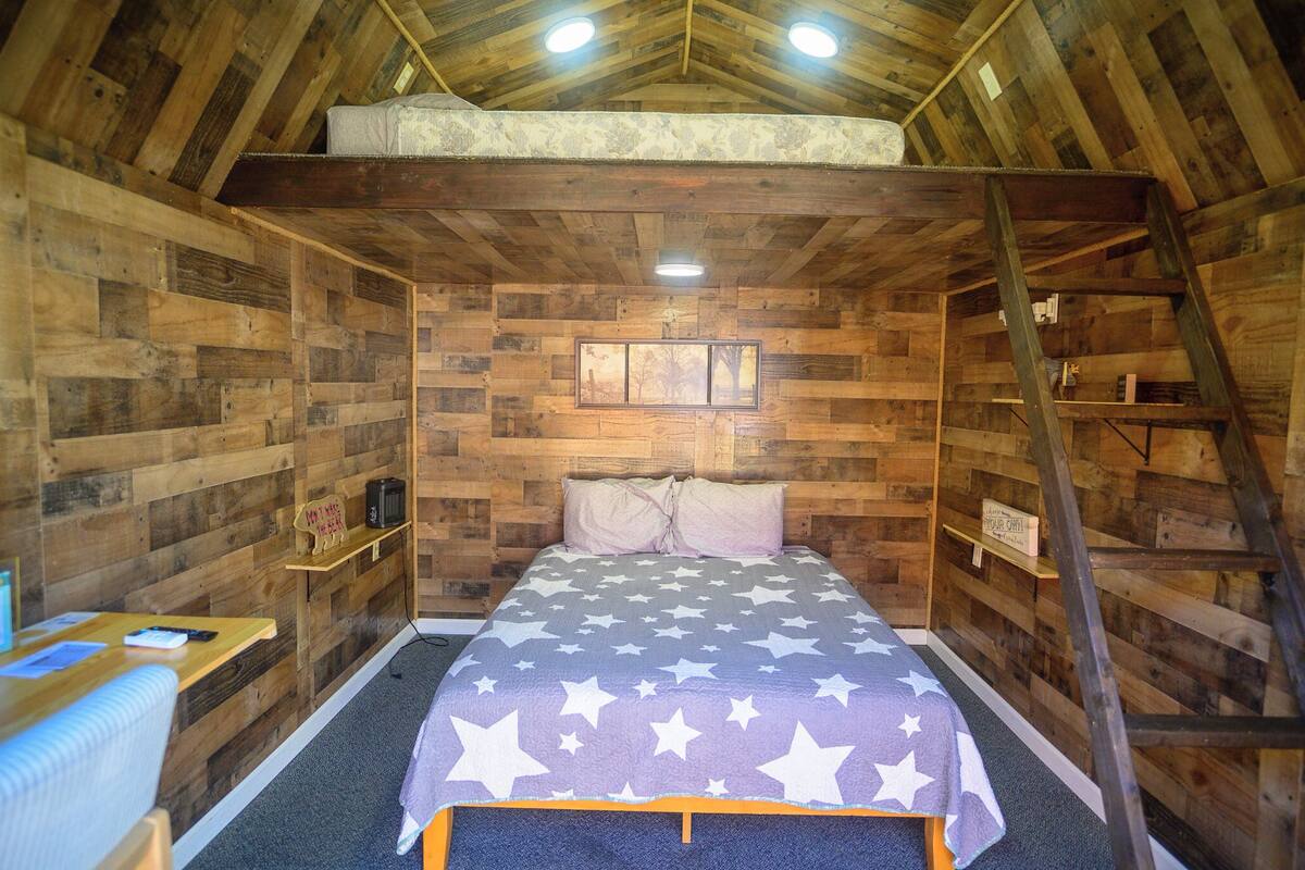 A wooden cabin room with a bed, a desk, and a ladder.