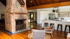 Amplia sala de estar con chimenea de ladrillo, piso de madera y cocina moderna con alacenas blancas y utensilios colgantes.