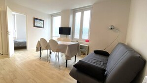 A living room with a sofa, a dining table with chairs, a TV, and a window with curtains. - Cozy Apartment with Free WiFi, Terrace, Near Trestraou Beach in Perros-Guirec (Perros-Guirec)