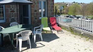 An outdoor seating area with a table and chairs, a stone building, and a view of parked cars and trees. - Cozy Apartment with Free WiFi, Terrace, Near Trestraou Beach in Perros-Guirec (Perros-Guirec)