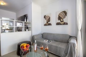 A modern living room with a grey sofa, a glass coffee table with a fruit basket and wine glasses, and artwork on the walls.