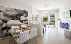A modern dining area with a glass table, white chairs, and a breakfast setting. There is a TV on the right and a balcony with a view.
