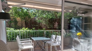Varanda com mesa, cadeiras, um vaso de planta e uma porta de vidro que dá para um jardim.