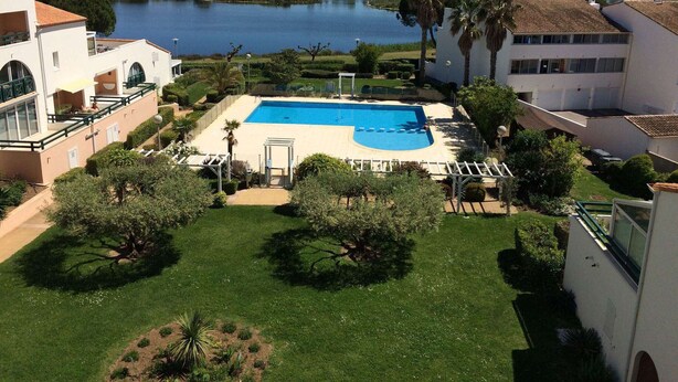 A swimming pool surrounded by greenery, a lake in the background, and buildings with balconies. - Duplex 3-room apartment with pool, air conditioning and garage in Port Camargue (Port Camargue)