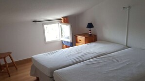 A small, simple room with two beds, a wooden chair, a nightstand with a lamp, and a window with curtains. - Duplex 3-room apartment with pool, air conditioning and garage in Port Camargue (Port Camargue)