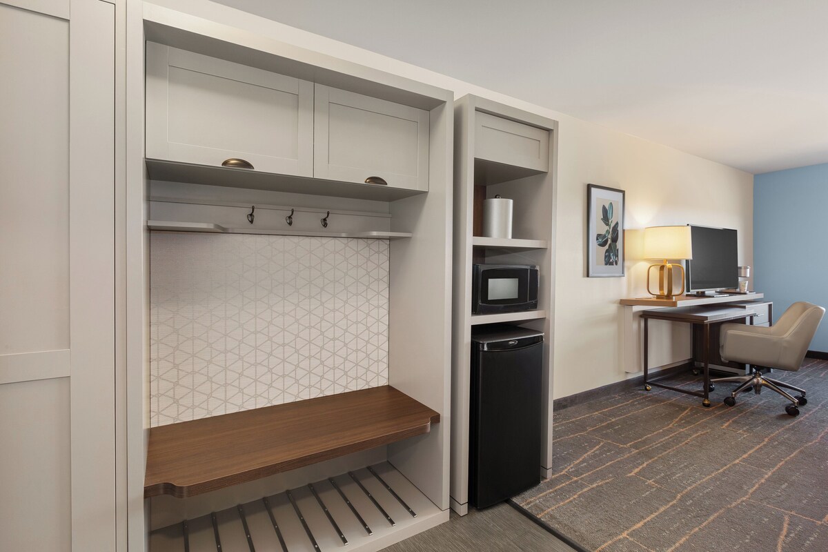 A compact hotel room with a kitchenette, a desk with a computer, a chair, and a small seating area.
