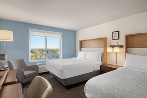 Habitación de hotel con dos camas, un escritorio, una silla, una ventana con vistas y una mesita de noche con lámparas.