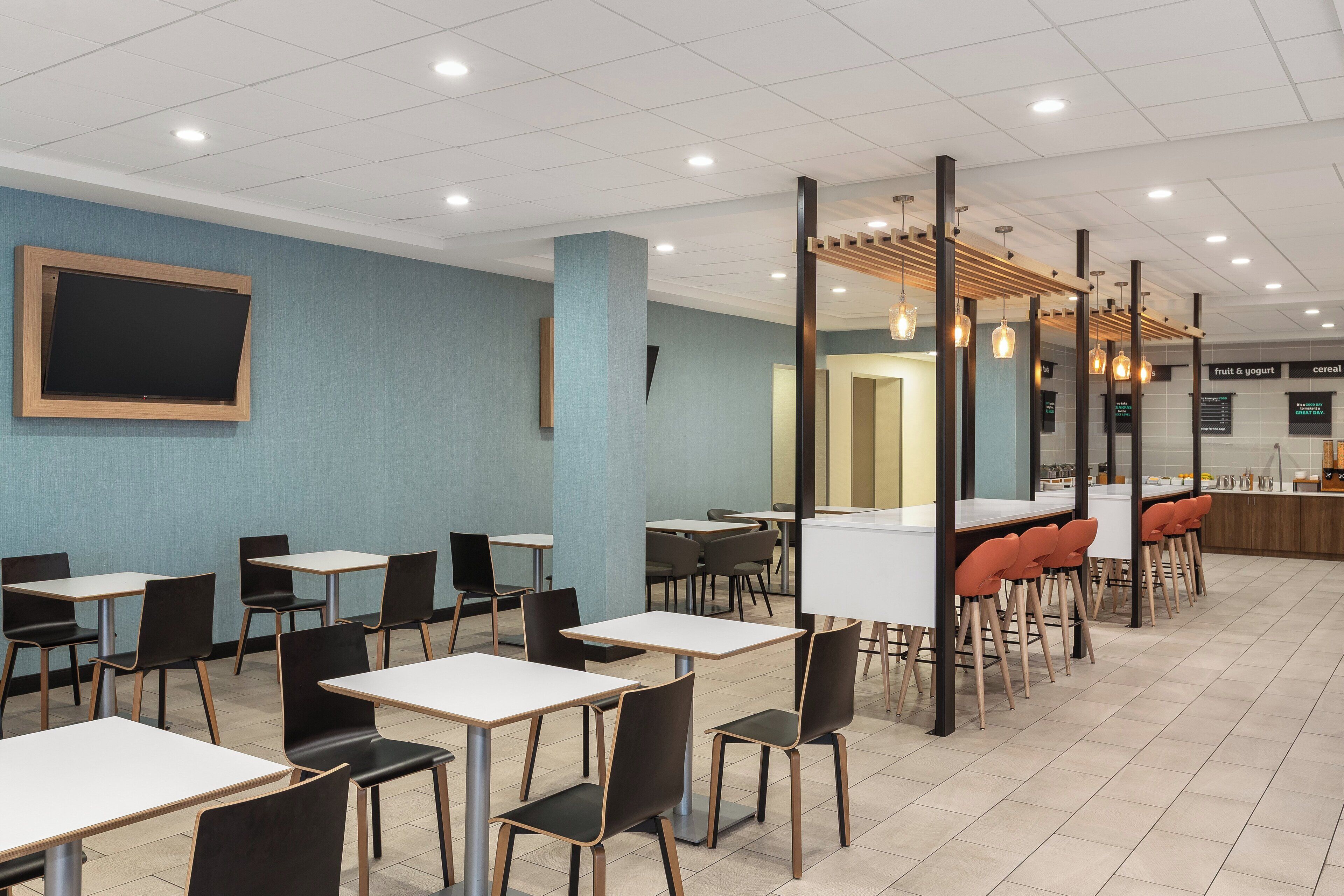 A cafeteria with tables and chairs, a large screen, and a menu board.