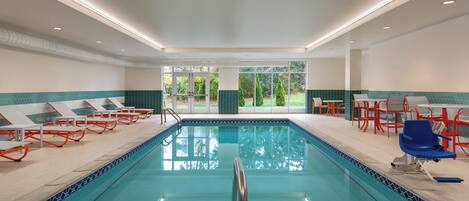 A swimming pool with lounge chairs and tables, surrounded by a tiled deck and a view of greenery.
