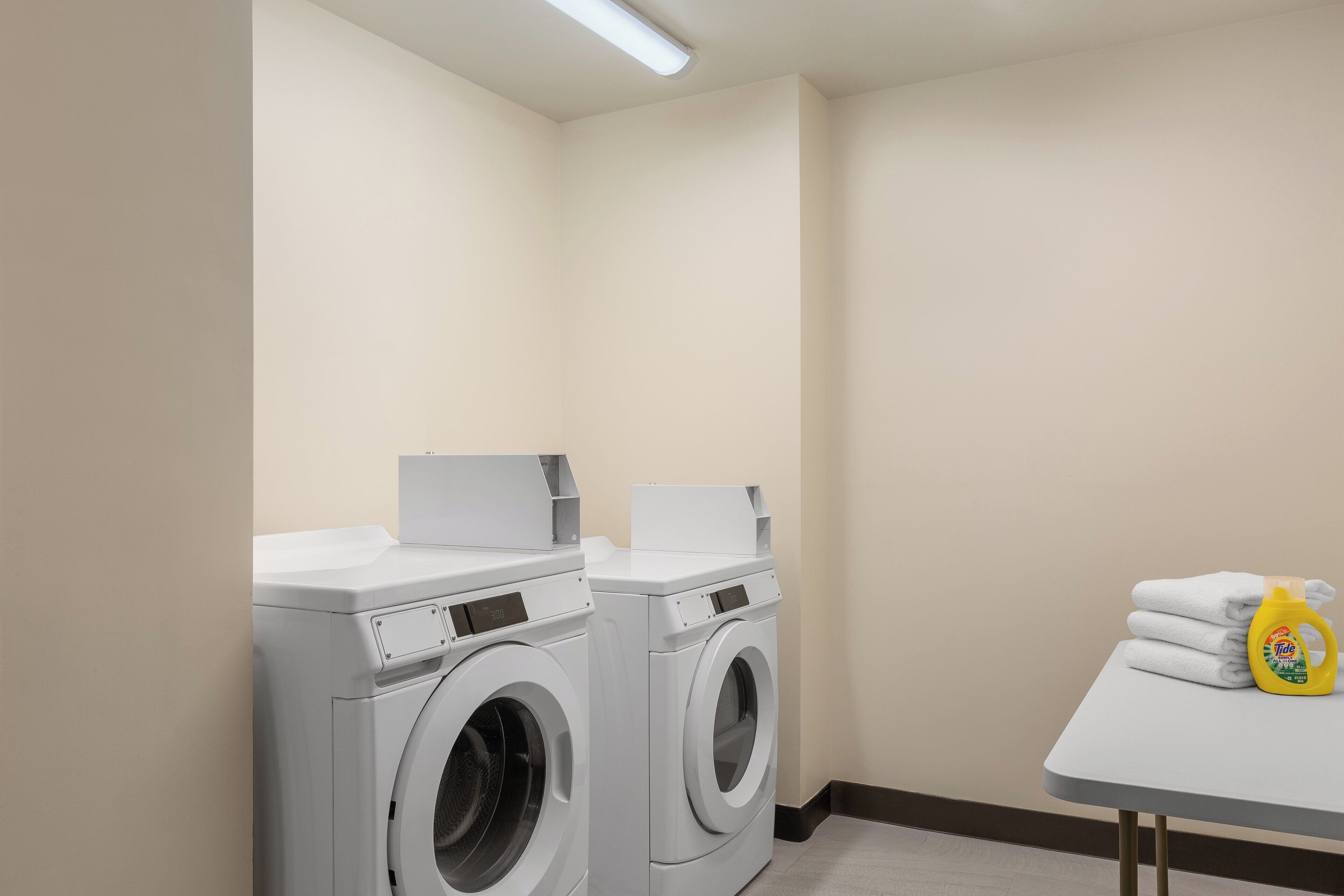 Una sala de bugaderia amb dues rentadores de càrrega frontal i una taula amb tovalloles pliegades i una ampolla de detergent.