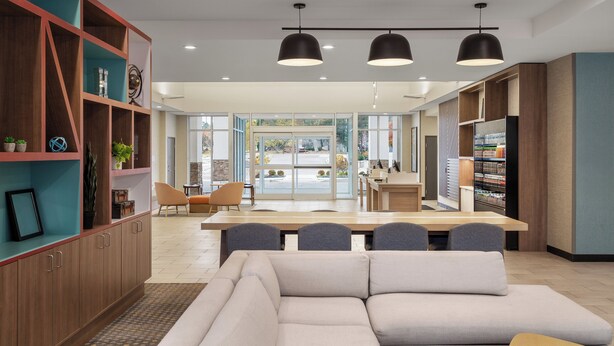 A modern interior with a sofa, dining table, and chairs in the foreground, and a kitchen area with a refrigerator and cabinets in the background. - Holiday Inn & Suites Detroit - Troy by IHG (Troy)