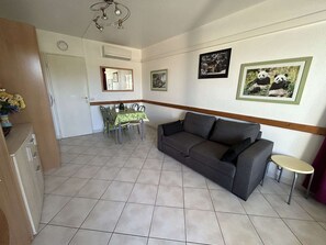 A living room with a sofa, a dining table with chairs, and a small round table. There are framed pictures on the wall, a clock, and an air conditioning unit.