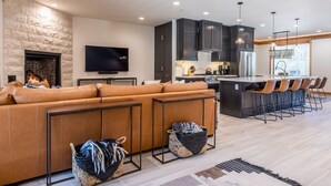 A modern living room with a large L-shaped sofa, a flat-screen TV, a fireplace, and a kitchen area with dark cabinets and a bar. - Luxury Mammoth Townhome w/ Hot Tub and Fire Pit (Mammoth Lakes)