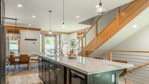 A modern kitchen with a large island, wooden staircase, and a dining area with a view of the outdoors. - Luxury Mammoth Townhome w/ Hot Tub and Fire Pit (Mammoth Lakes)