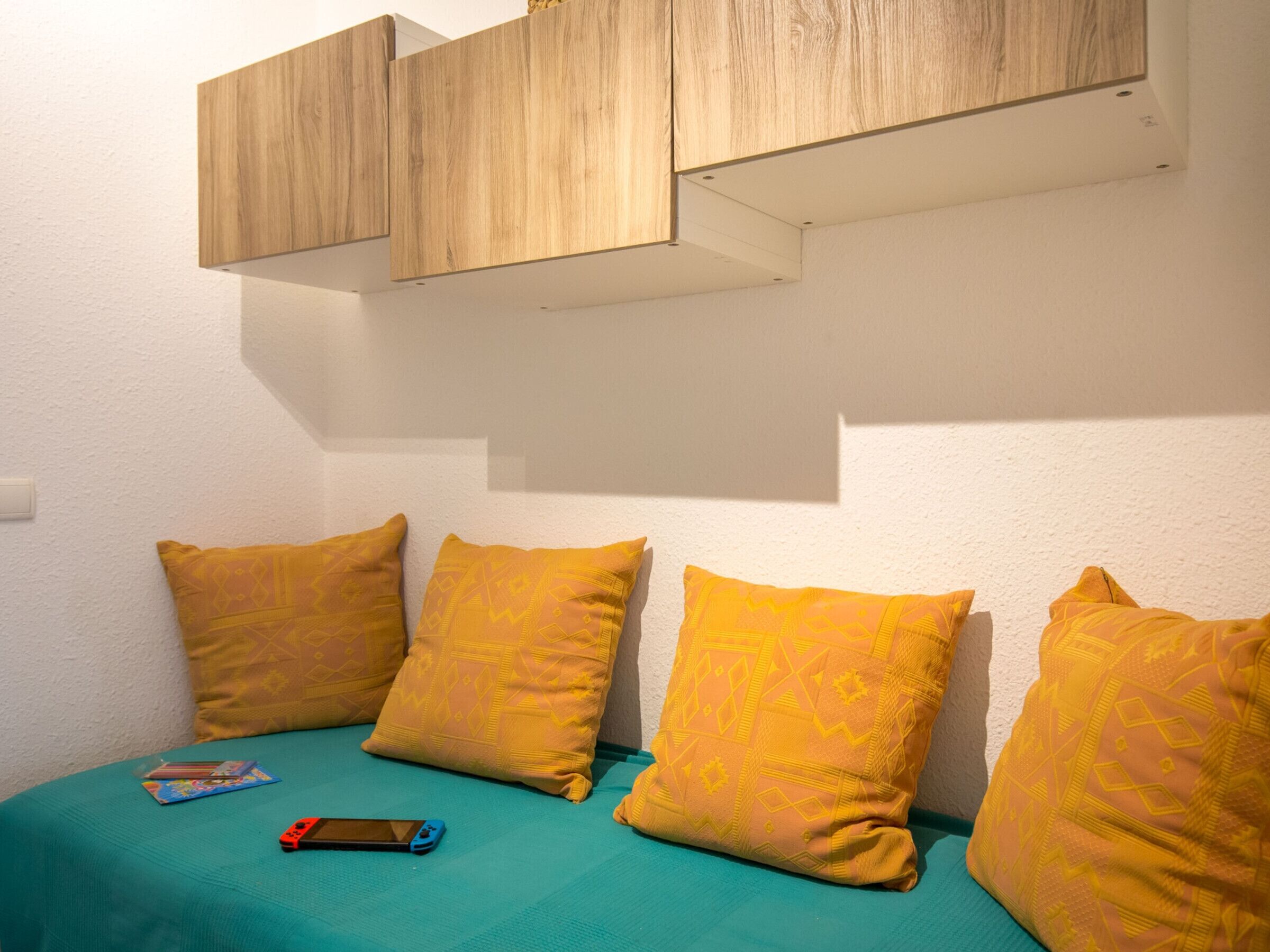A room with a bed, three yellow pillows, a teal bedspread, and a wooden cabinet above the bed.