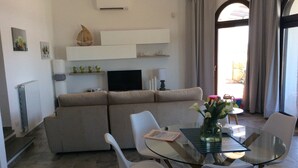 A modern living room with a beige sofa, a glass coffee table, and white chairs. There is a flat-screen TV mounted on the wall, a small shelf with decorative items, and a window with curtains. - 3 Bedroom Villa in Alghero (Alghero)