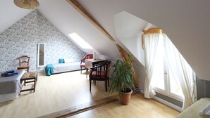 Ruime zolderkamer met een schuine kap, een houten tafel, stoelen, een potplant en een groot raam met gordijnen.