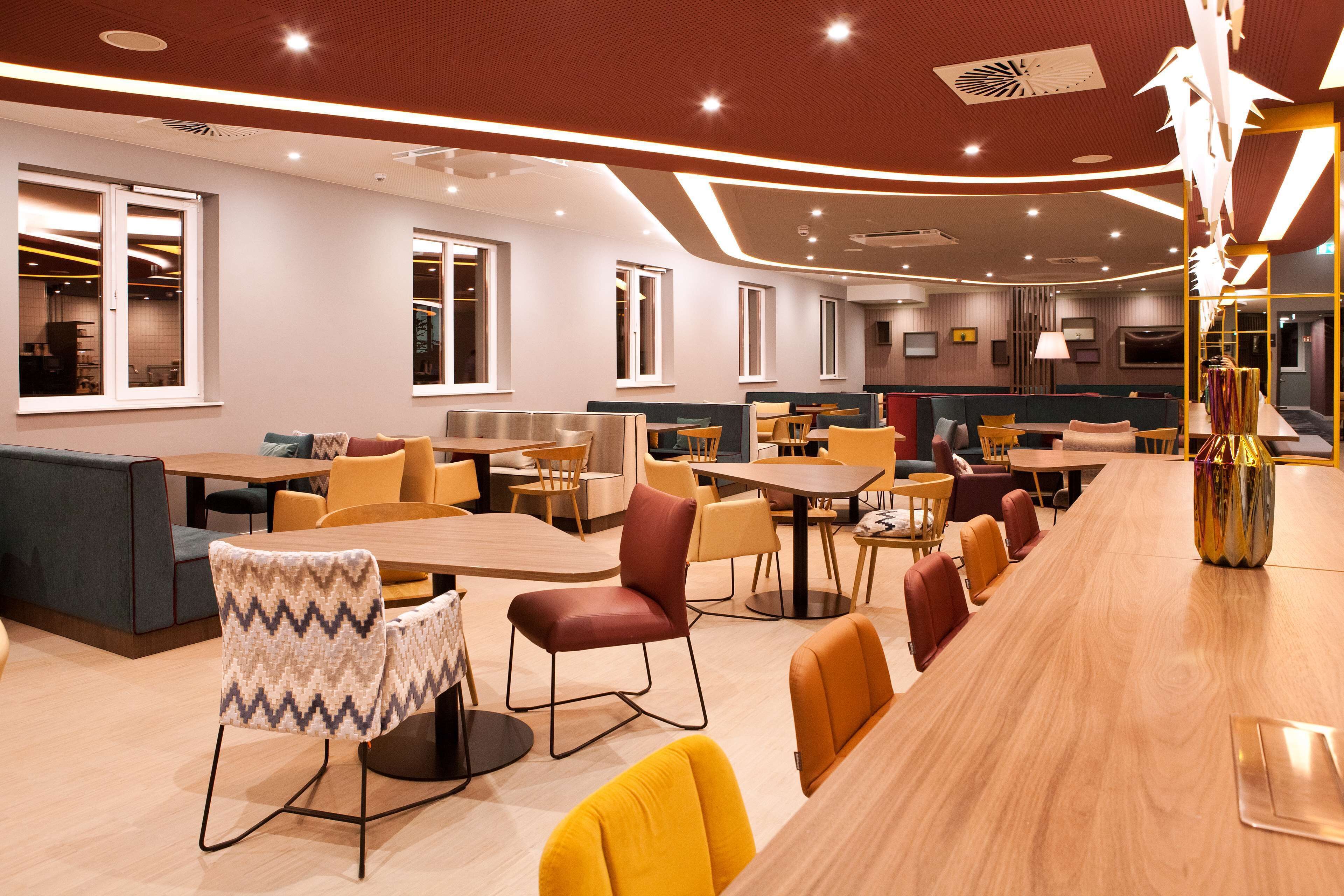 A modern restaurant with a long wooden bar, multiple seating areas including booths and tables, and large windows providing natural light.