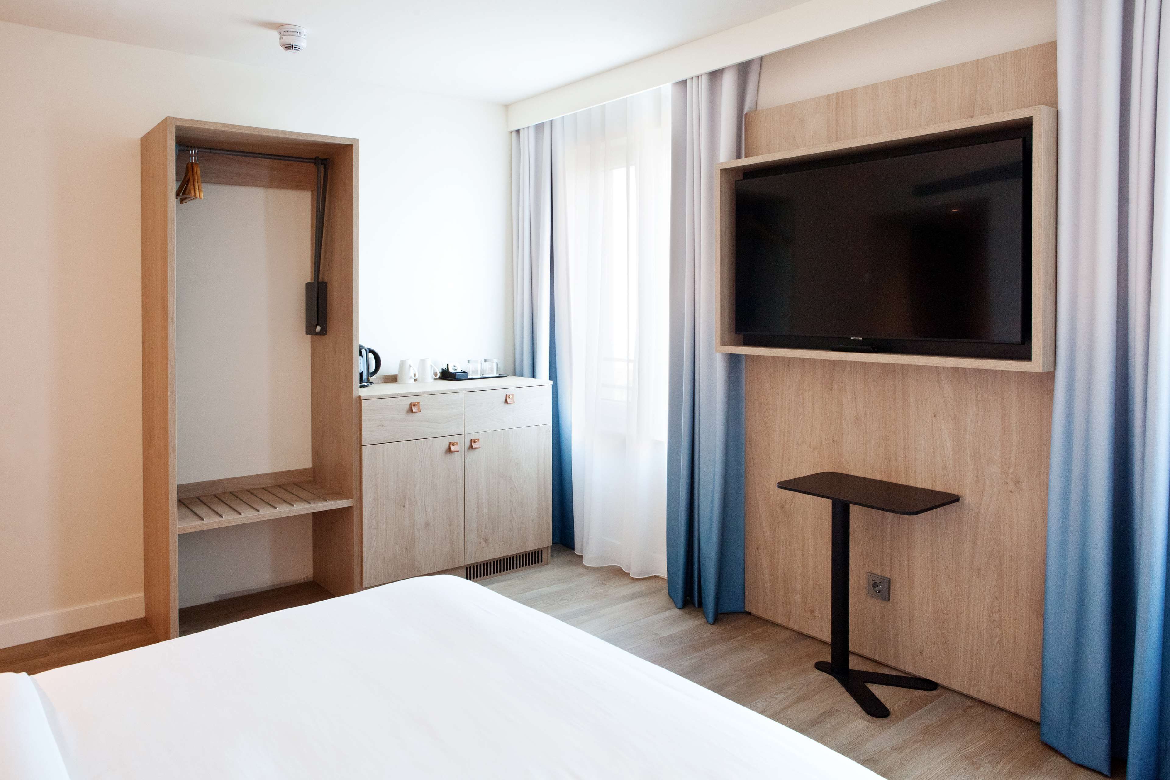 Habitación de hotel con cama, televisor montado en la pared, armario de madera, una mesita y una ventana con cortinas.