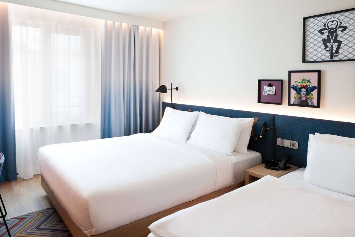 Ein Hotelzimmer mit zwei Betten, einem Nachttisch mit Lampe und einem Fenster mit durchsichtigen Vorhängen.