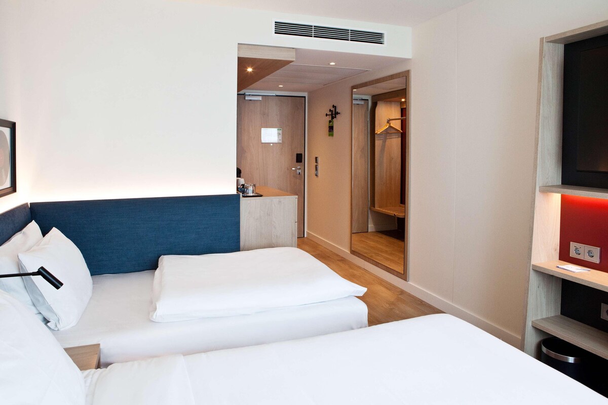 A hotel room with two beds, a wooden headboard, a small desk, a mirror, a TV, and a red-lit shelf.