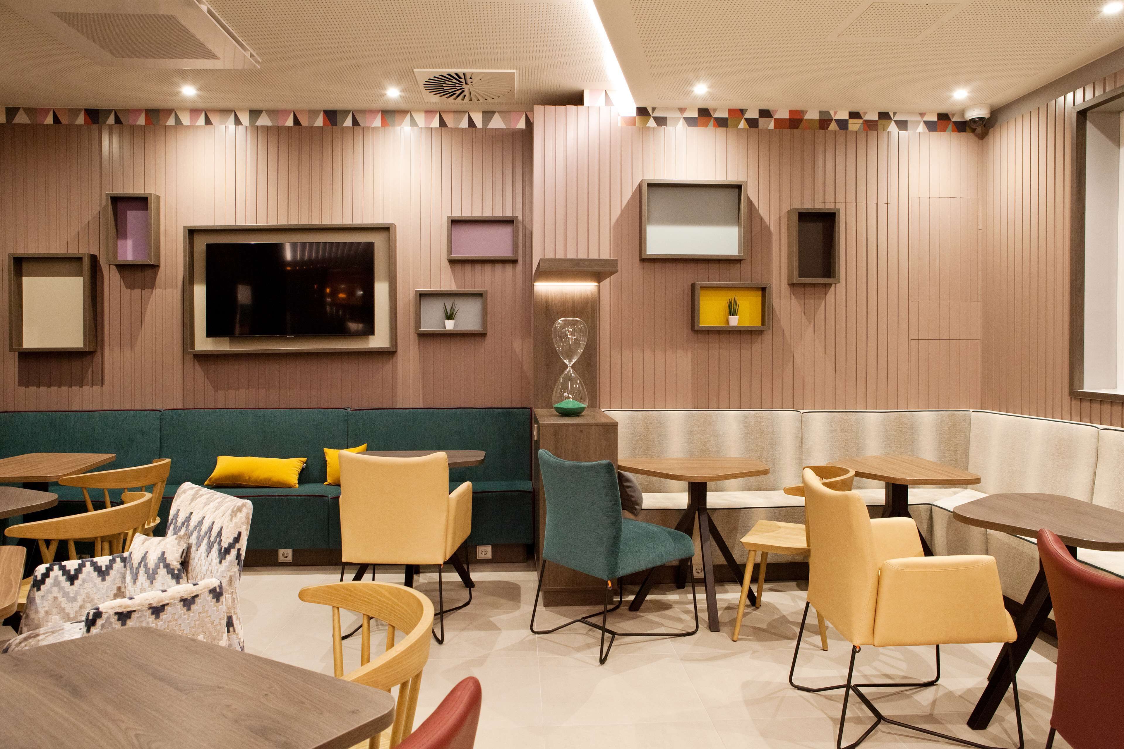 Een modern interieur met houten lambrisering, een groene bank, gele en teal stoelen, en een grote flatscreen televisie.