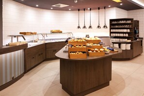 Une cuisine moderne dotée d’un îlot central où sont exposés des pâtisseries, ainsi que d’un coin café et d’une étagère où sont rangés avec soin des verres.
