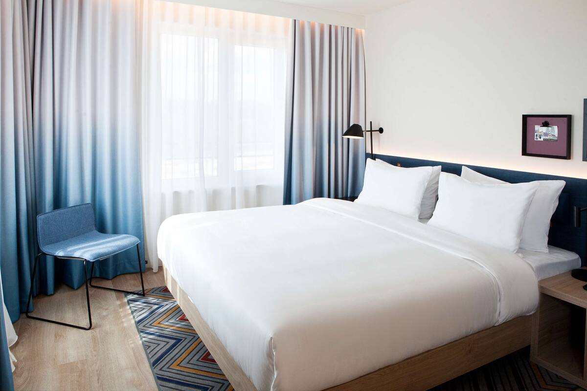 Ein Hotelzimmer mit einem großen Bett, einem Sessel, einem Schreibtisch und einem Fenster mit Vorhängen.