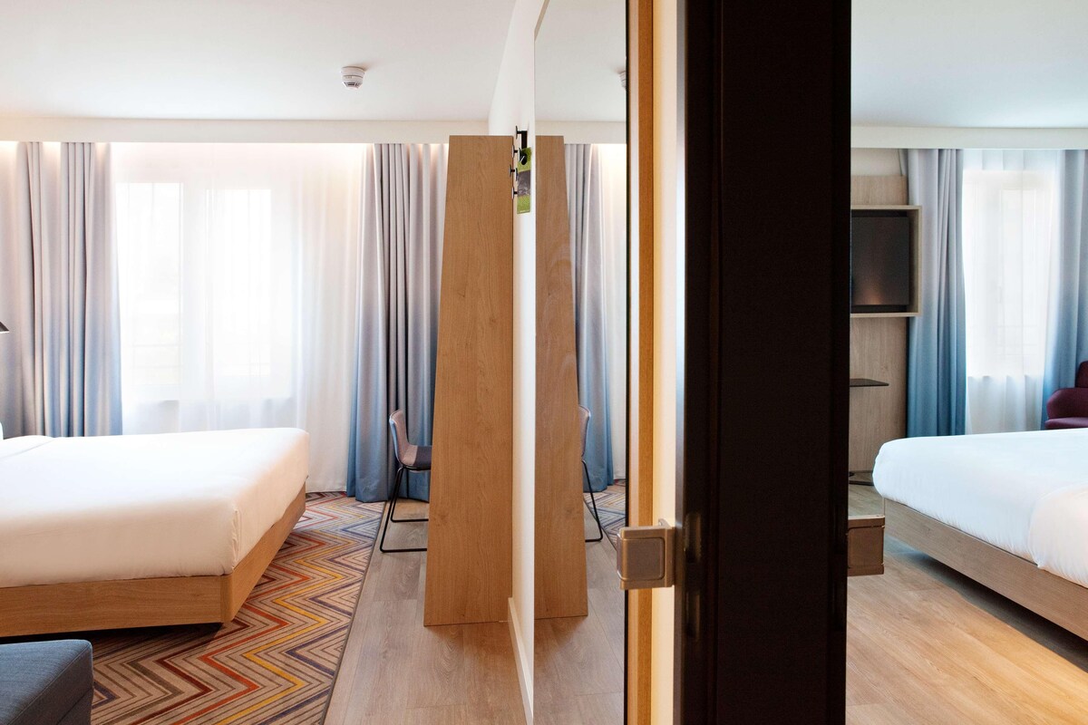 A hotel room with two beds, a wooden wardrobe, a chair, and a patterned rug.