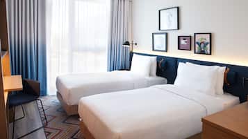 Habitación de hotel con dos camas, un escritorio, una silla y un ventanal grande con cortinas.