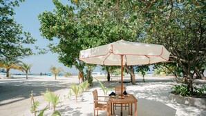 Une table installée sur la plage, protégée par un parasol blanc, entourée de sable et de verdure.