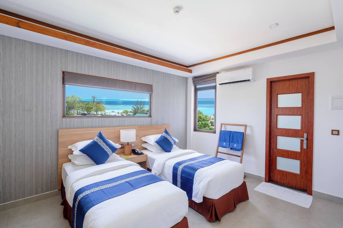 A hotel room with two beds, a large window with a view of the sea, a wooden door, and an air conditioning unit.