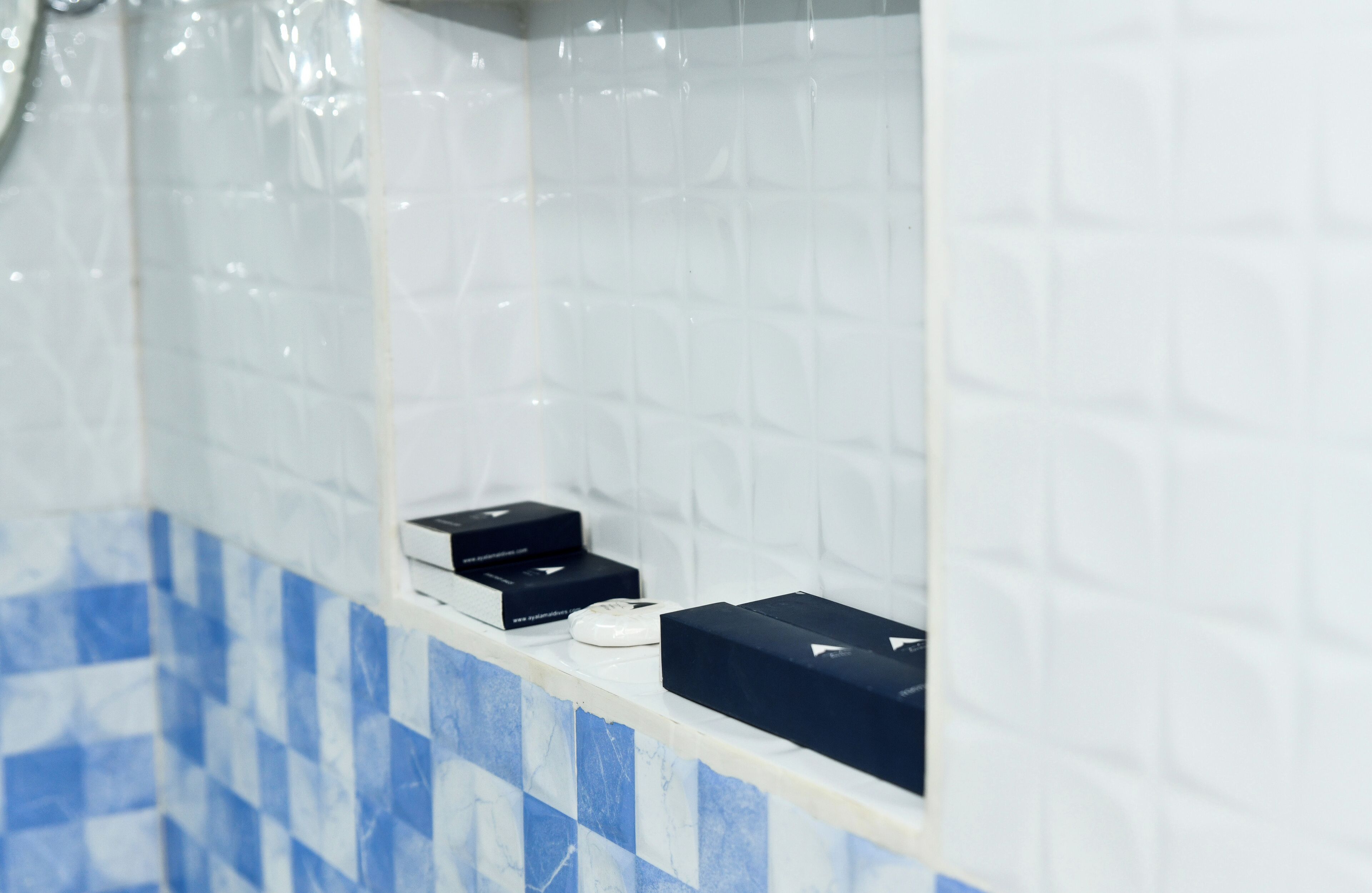 Une salle de bain avec des murs recouverts de carreaux blancs et un sol à carreaux bleus et blancs. On peut voir deux boîtes noires et un objet blanc sur le comptoir.