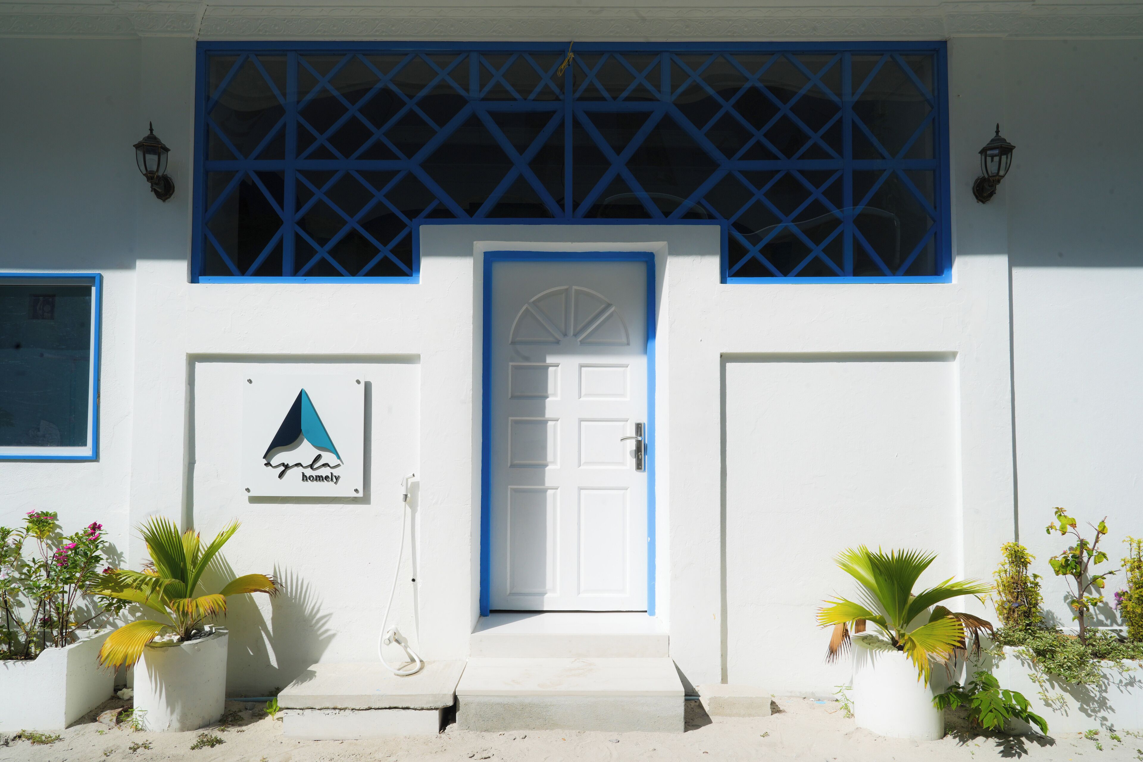 A white building with a blue door and window frames, a sign with a mountain and tent icon, and potted plants at the entrance.