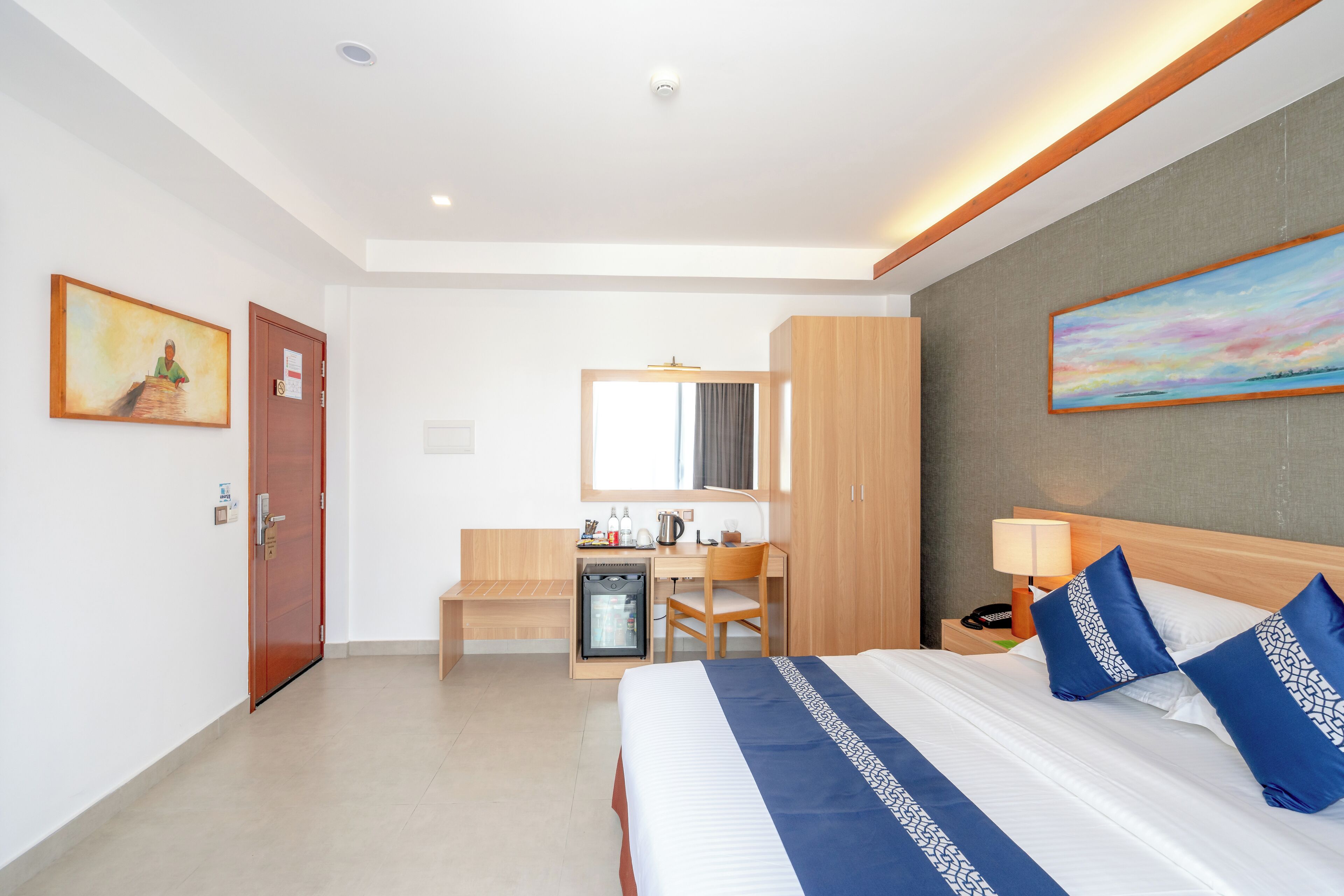 A hotel room with a bed, a desk, a mini-fridge, a chair, and a nightstand with a lamp.