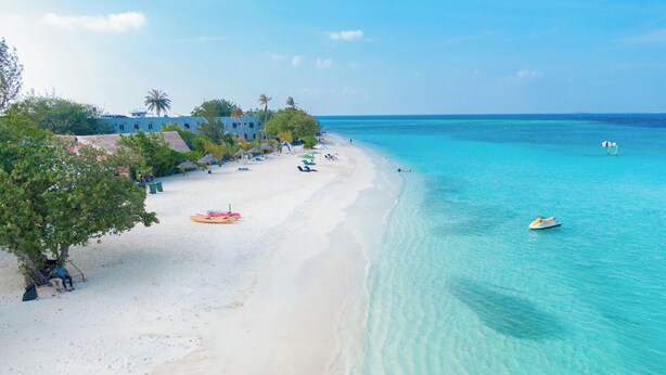 Une plage avec une eau turquoise et limpide, un rivage sablonneux et quelques personnes qui se promènent. On aperçoit des parasols et un bateau.