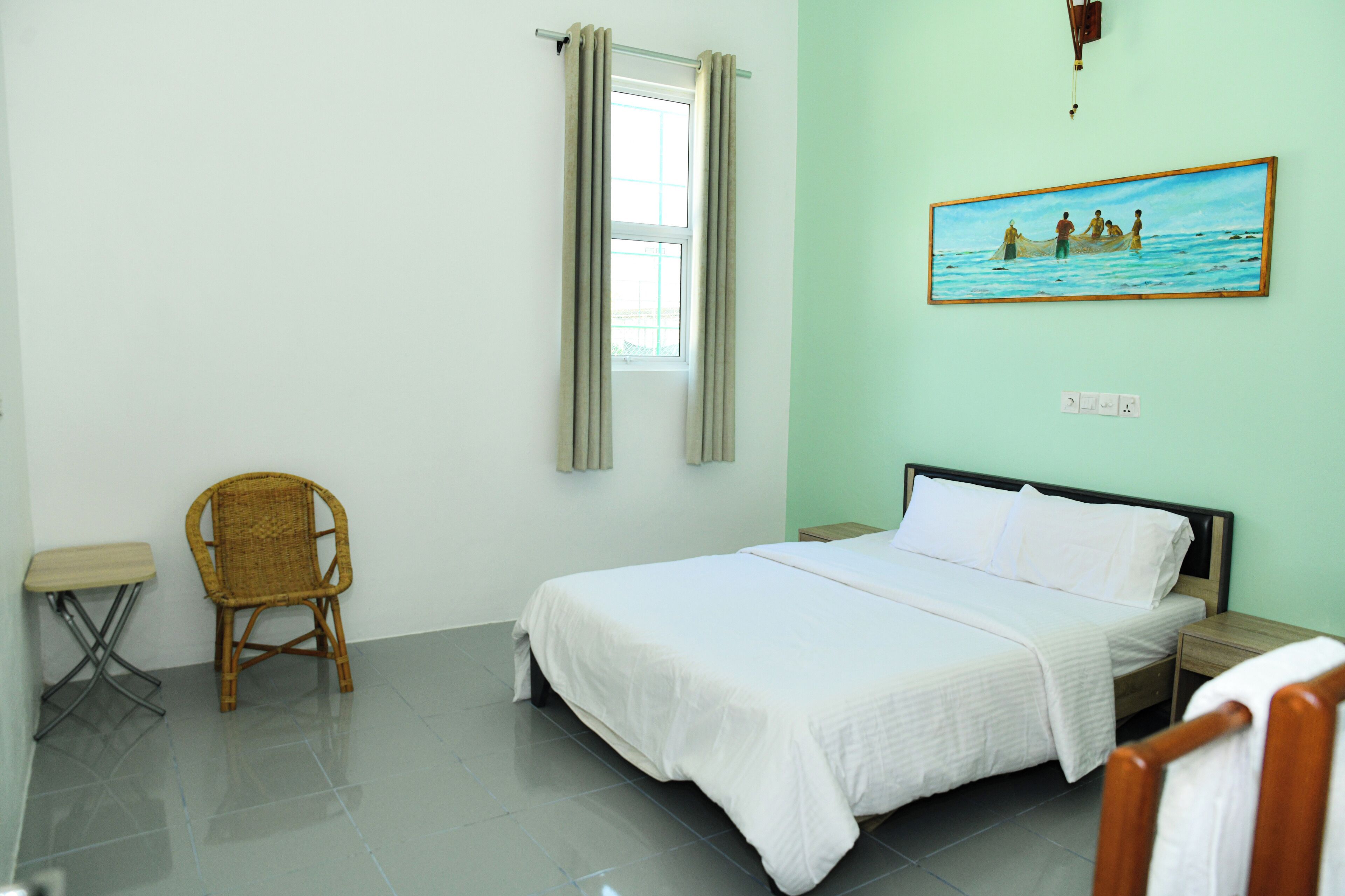 A hotel room with a bed, a chair, a small table, a painting on the wall, and a window with curtains.