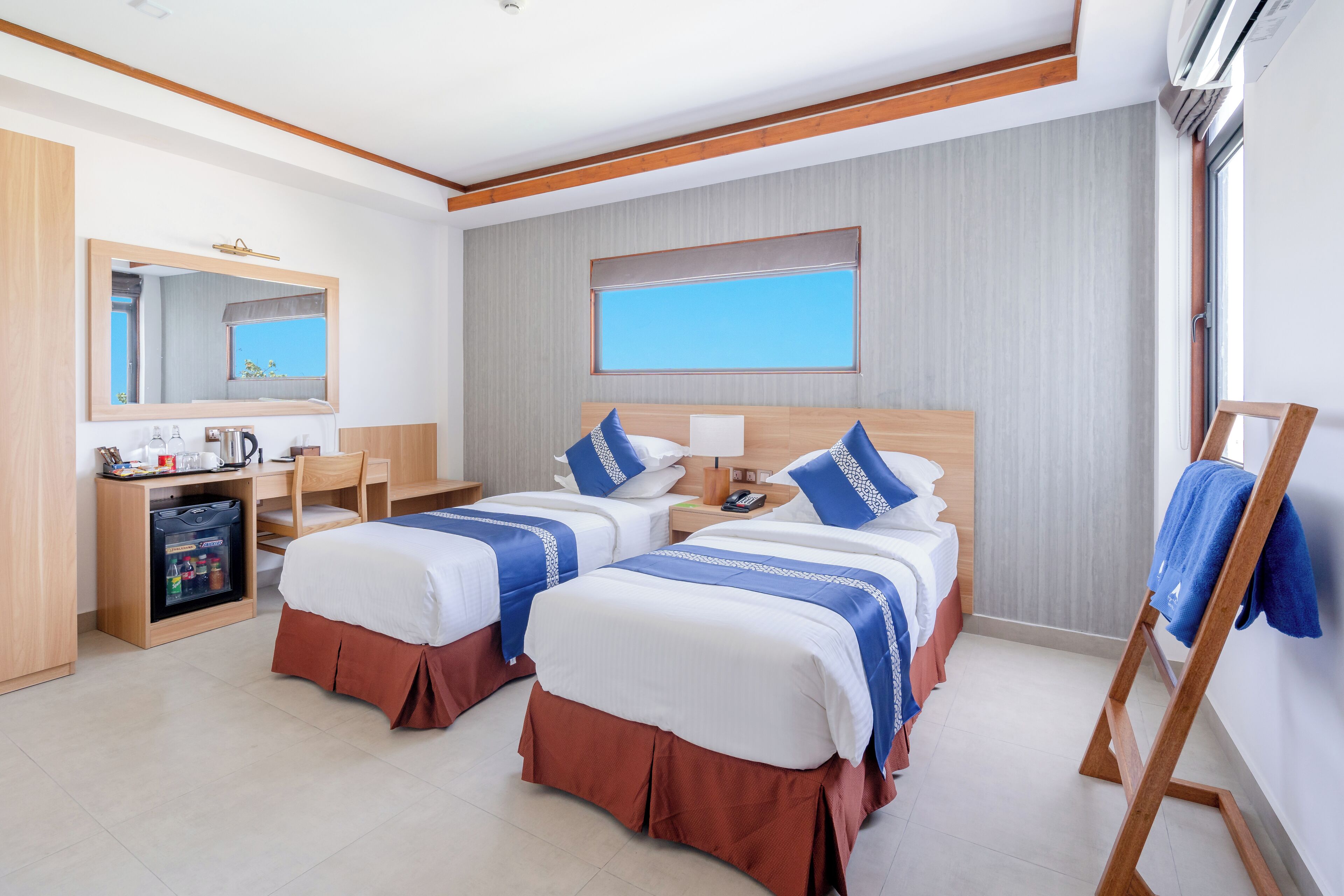 A hotel room with two beds, a desk, a minibar, and a large window.