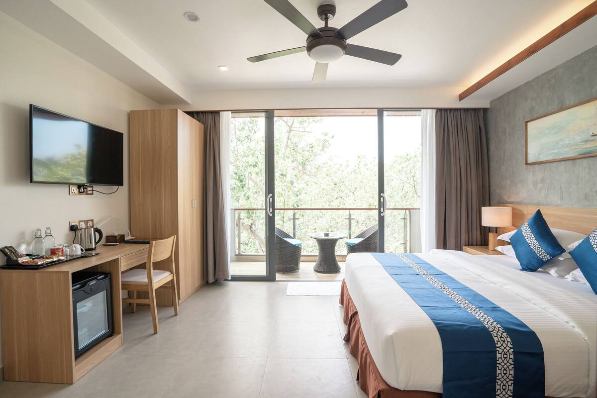 A hotel room with a bed, a desk with a television, a ceiling fan, and a balcony with a table and chairs.