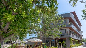 Un bâtiment moderne à la façade incurvée, doté d’un espace extérieur aménagé avec des tables et des chaises, ainsi que de diverses plantes et arbres.