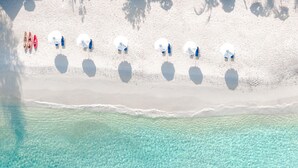 Vue aérienne d’une plage aux eaux turquoise et limpides, au sable blanc, où l’on aperçoit plusieurs parasols et bateaux.