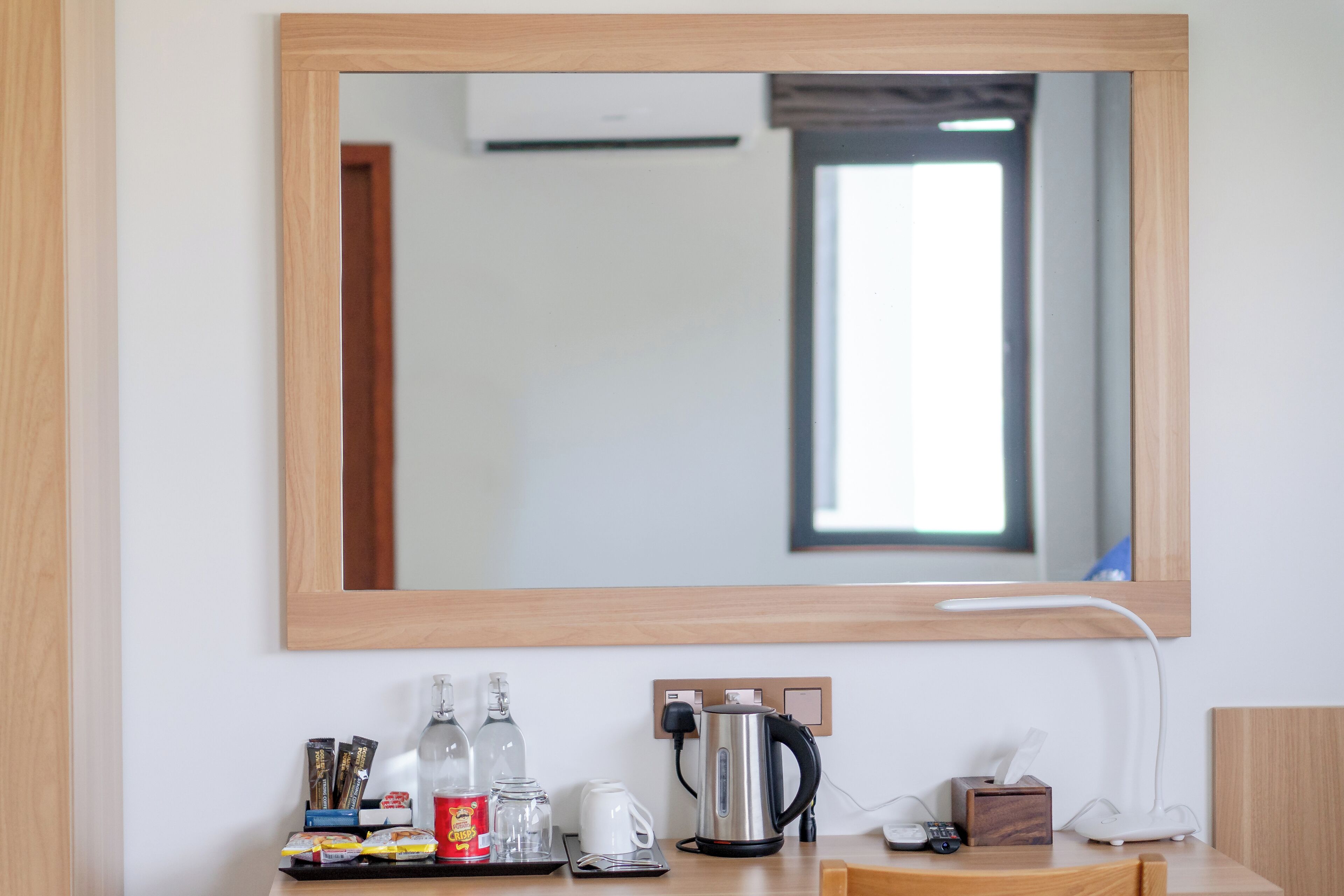 A hotel room with a wooden-framed mirror, a coffee maker, a kettle, a tray with snacks, and a tissue box.