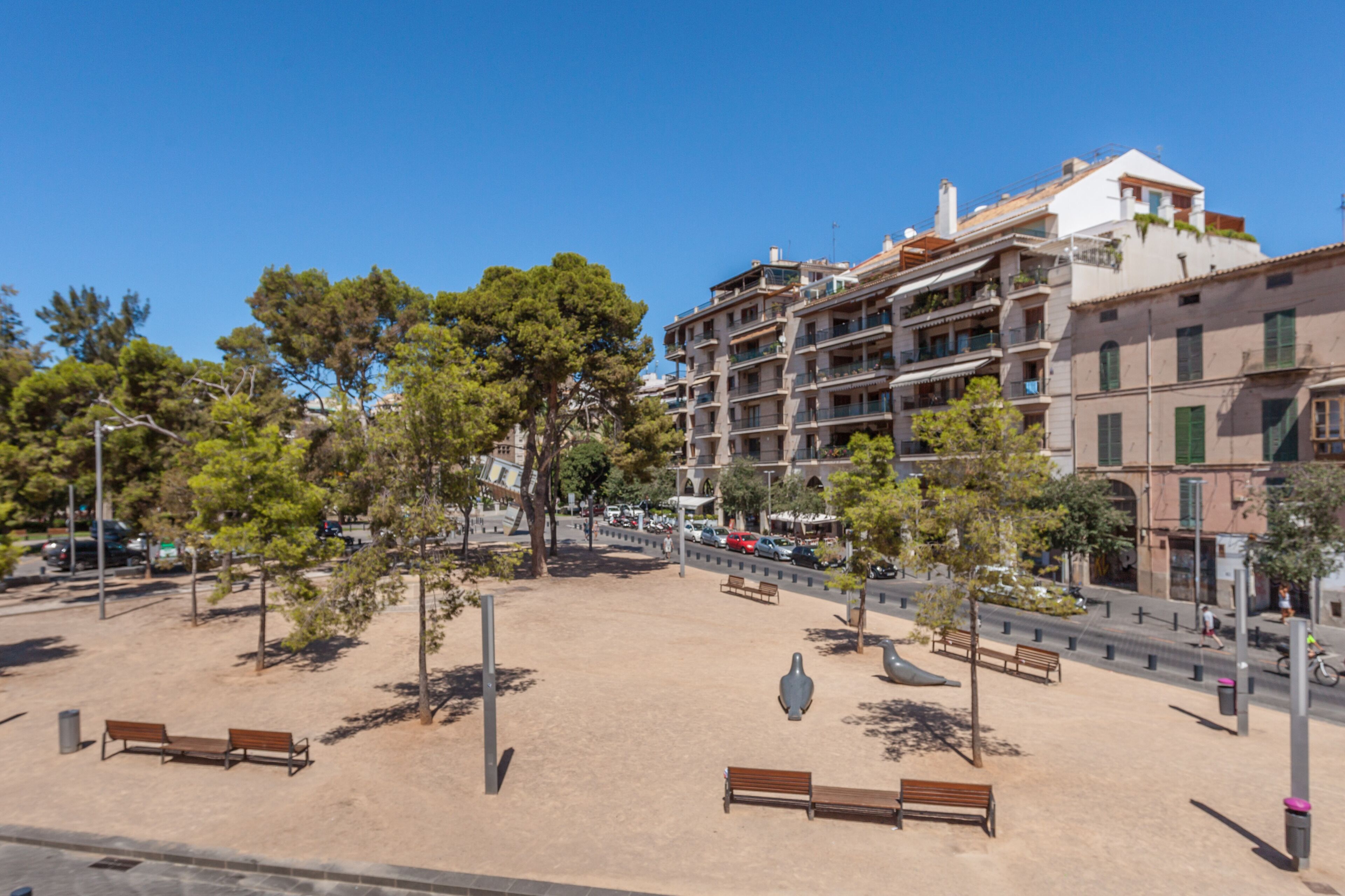 A public square with benches, trees, and a multi-story building with balconies.