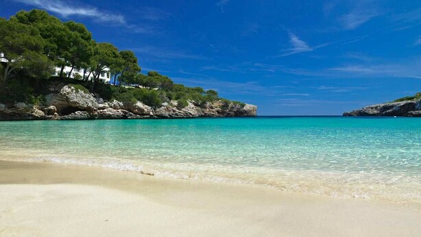 Ein Strand mit klarem türkisblauem Wasser, weißem Sand und einer felsigen Klippe mit Bäumen und Gebäuden.