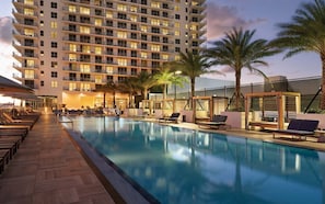 Ein Poolbereich eines Hotels mit großem Pool, Liegestühlen und einem Hochhaus im Hintergrund.