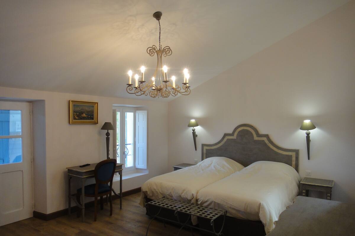 A bedroom with a double bed, a desk with a chair, a standing lamp, and a chandelier.