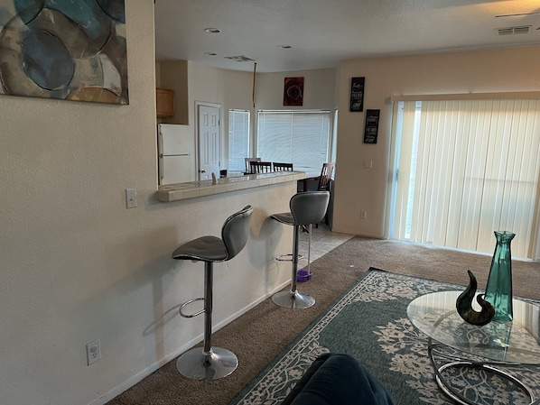 A modern living room with a glass coffee table, two bar stools, a patterned rug, and a view of the kitchen through a glass door.