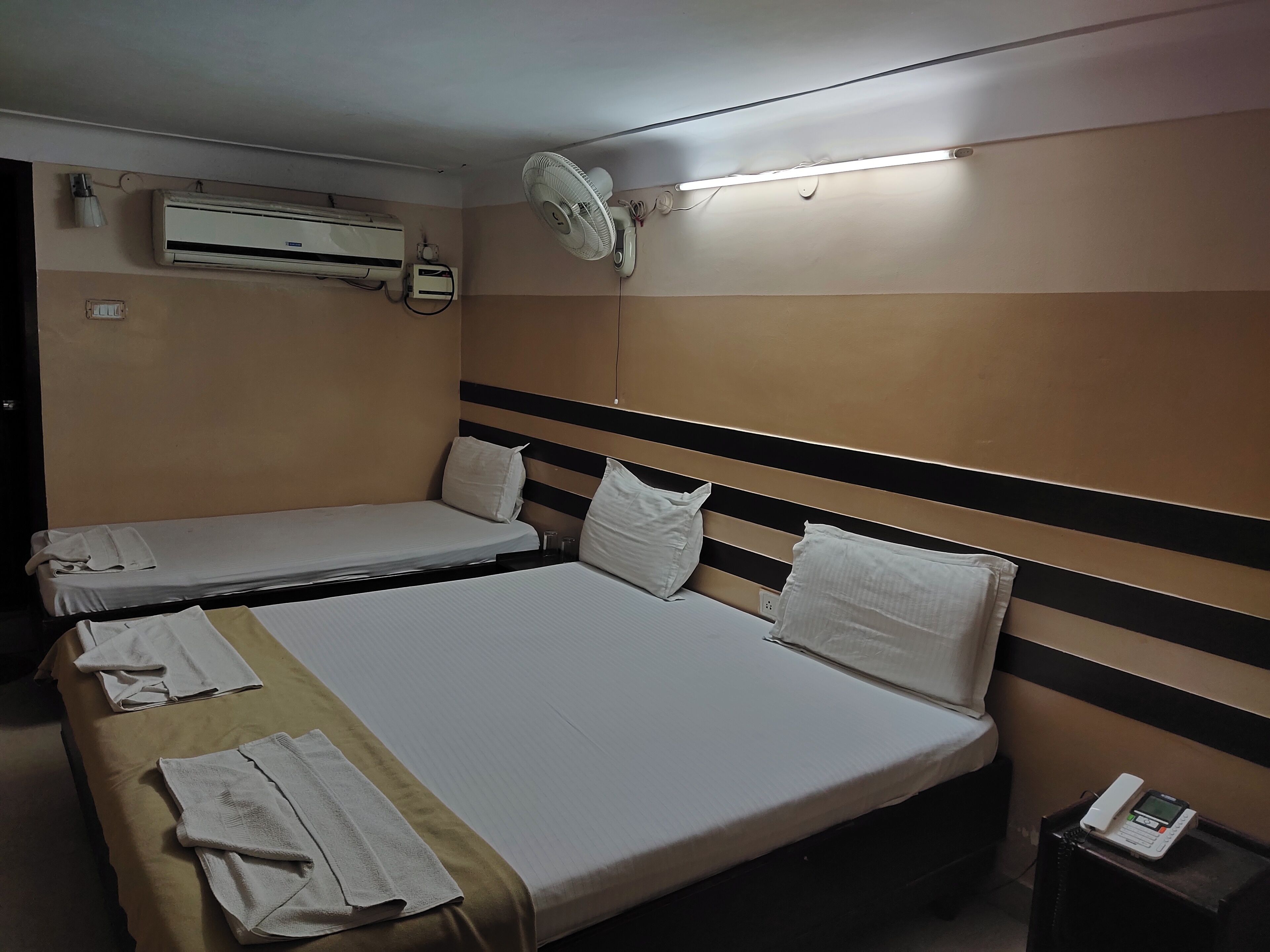 Une chambre d’hôtel avec deux lits, un climatiseur, un ventilateur de plafond et un téléphone sur la table de chevet.
