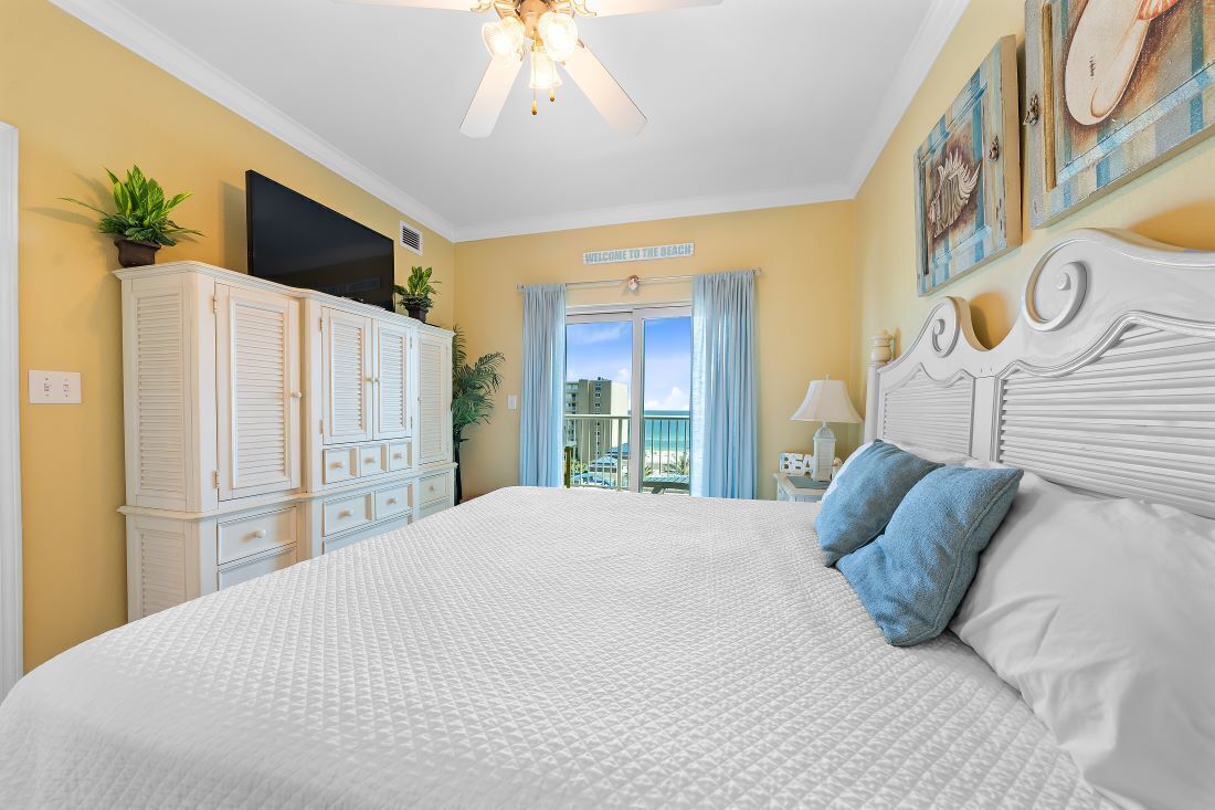 A bedroom with a large bed, a ceiling fan, a TV on a cabinet, and a view of the beach through a window.