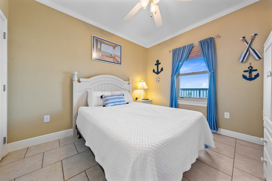 Une chambre à coucher comprenant un lit, un ventilateur de plafond, une fenêtre avec des rideaux et des éléments de décoration murale sur le thème de la marine.
